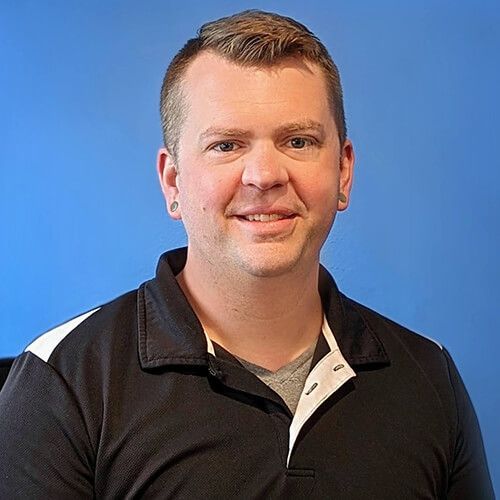 A man with short hair smiles at the camera, wearing a black polo shirt and grey t-shirt with a blue backdrop. | King's Auto Center