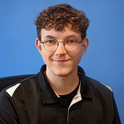 Young man with curly brown hair and glasses smiles at the camera; wears a black polo with white accents against a blue backdrop. | King's Auto Center