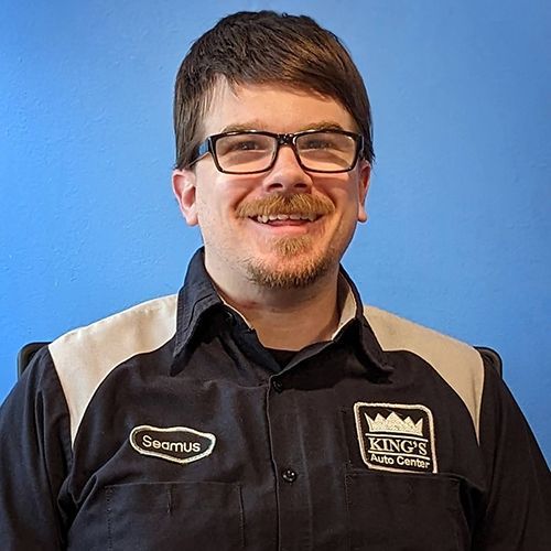 Man with glasses, wearing a mechanic's uniform with a King's Auto Center logo, smiles in front of a blue wall.