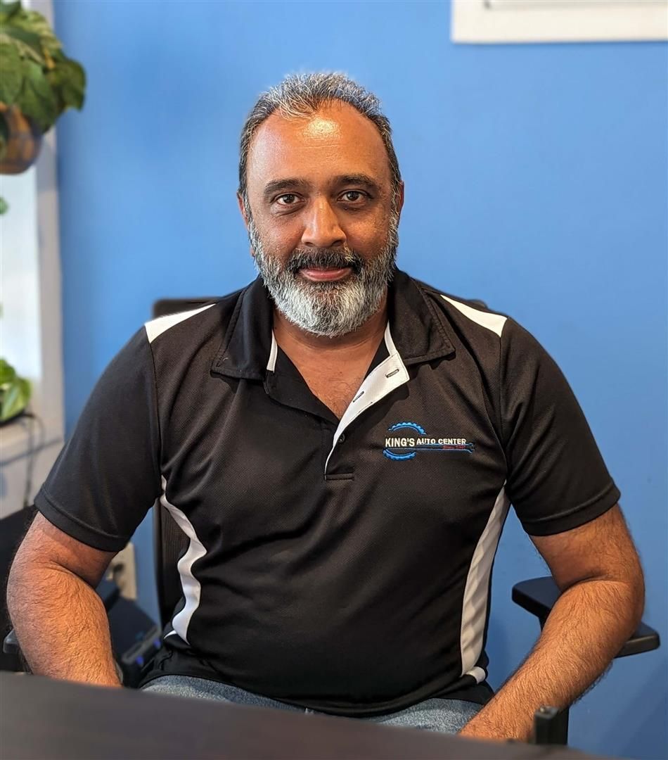 Man with a graying beard wearing a black polo shirt, smiling at the camera in an office setting with blue walls. | King's Auto Center