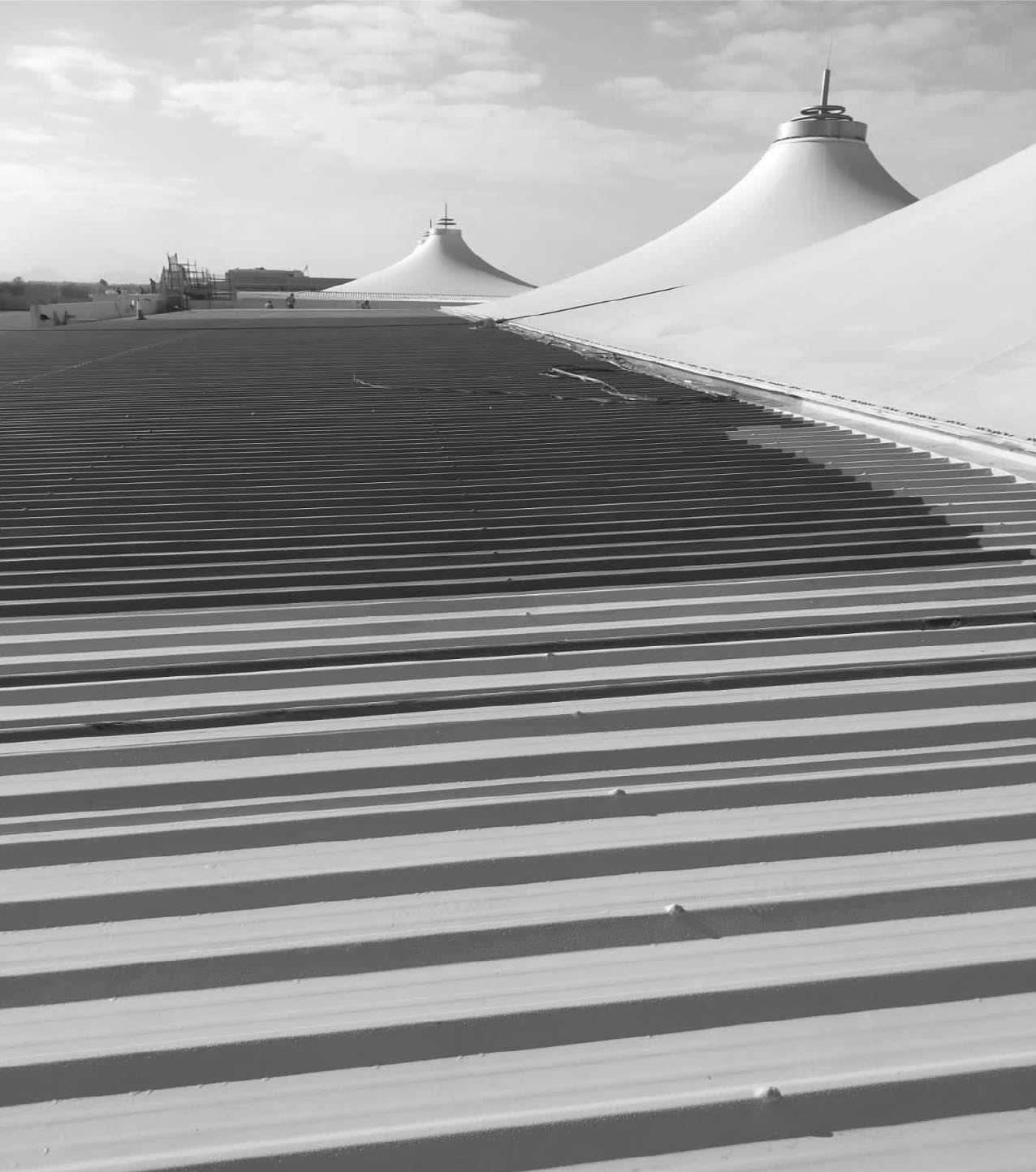 Black and white photo of a large, modern building with white tent-like structures and a corrugated roof casting long shadows.