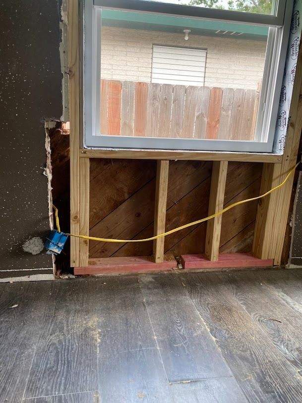 Partially gutted wall beneath a window with exposed studs and a yellow cord in an unfinished room