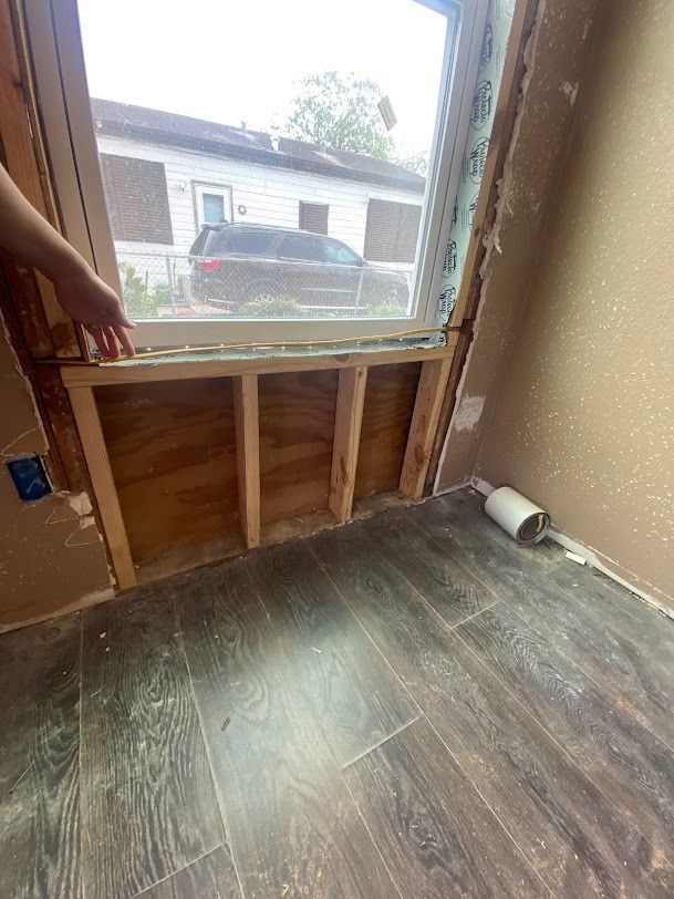Partially stripped wall with a window opening and exposed studs in a room with wood flooring