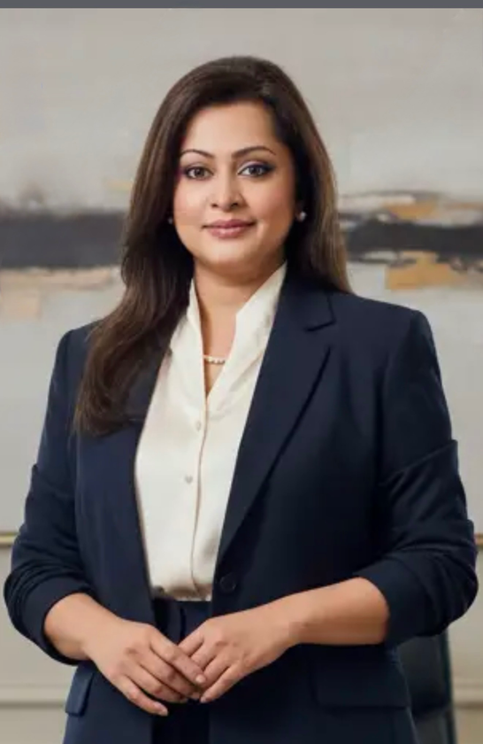 Portrait of a woman in a navy blazer and white blouse, standing indoors against a blurred neutral background.