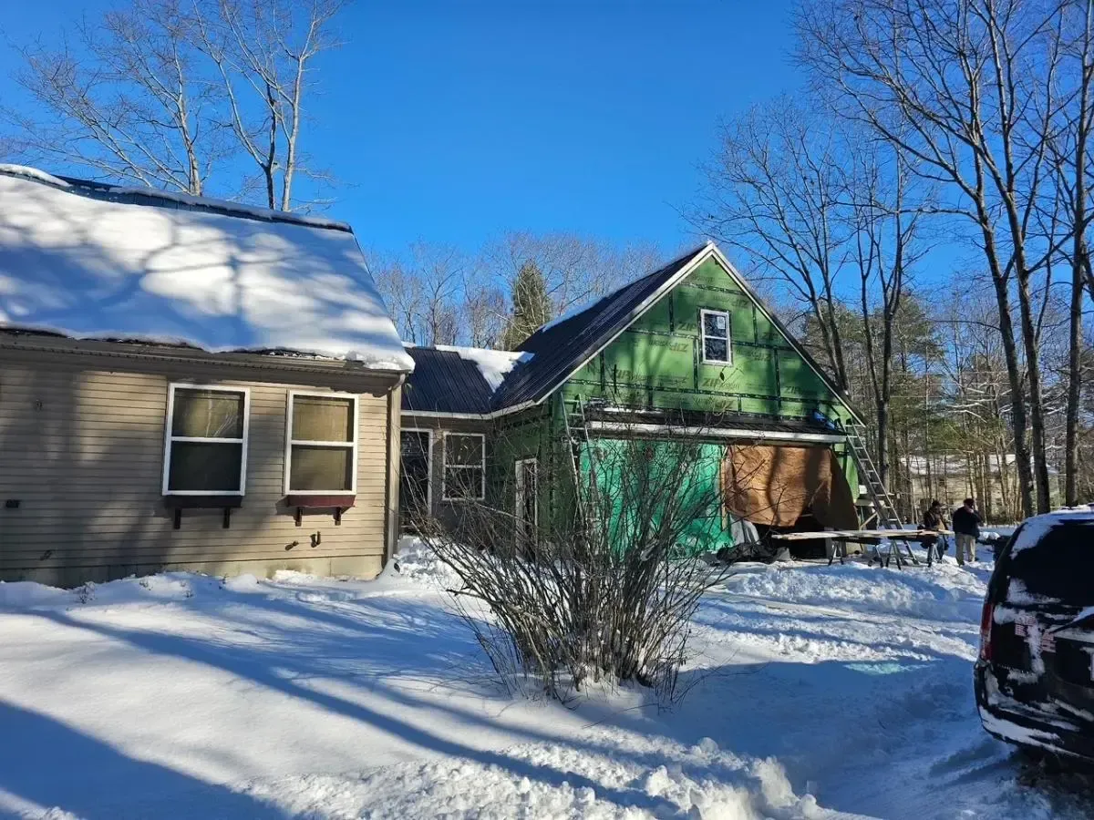 Snowy scene of two buildings; one under construction with green wrap, other beige, and bare trees.