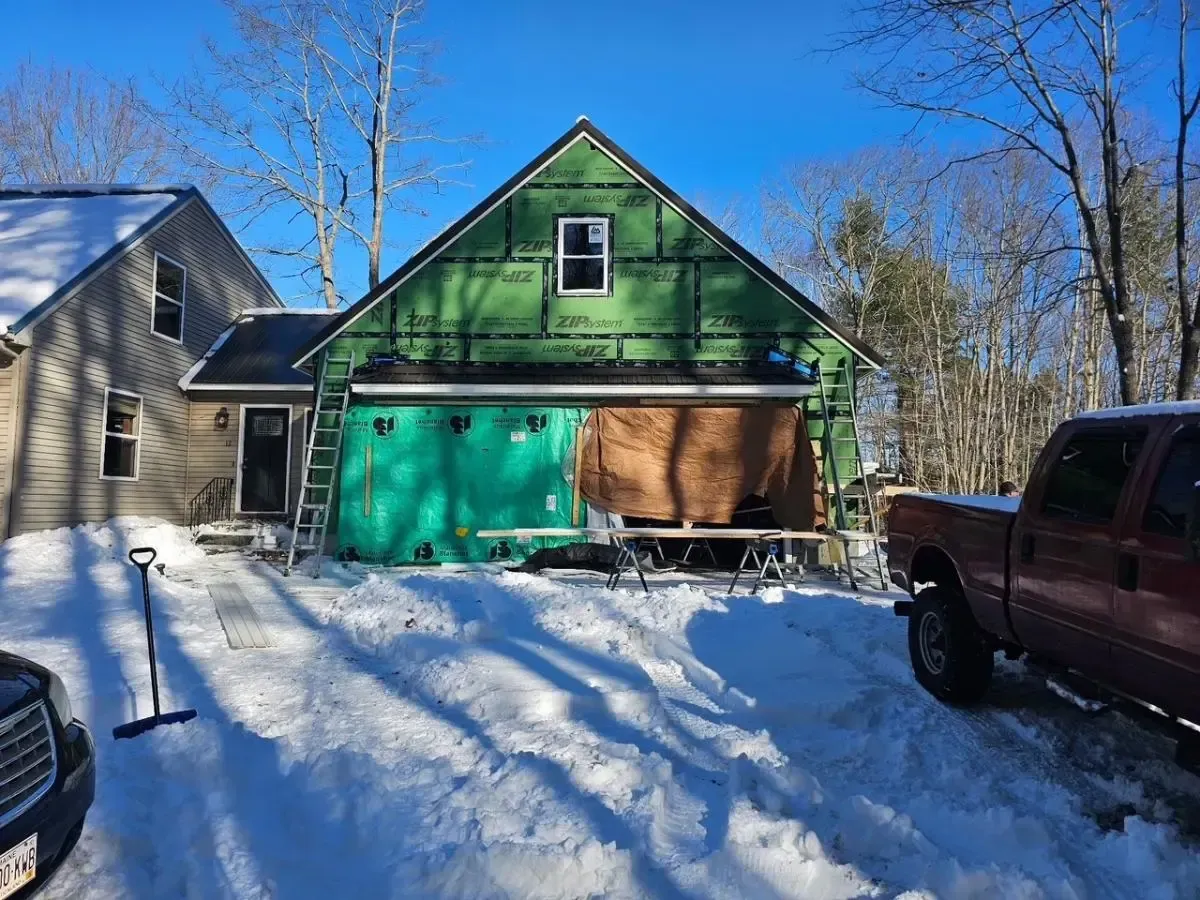 Construction in progress: house addition with green wrap and exposed framing, surrounded by snow; truck parked nearby.