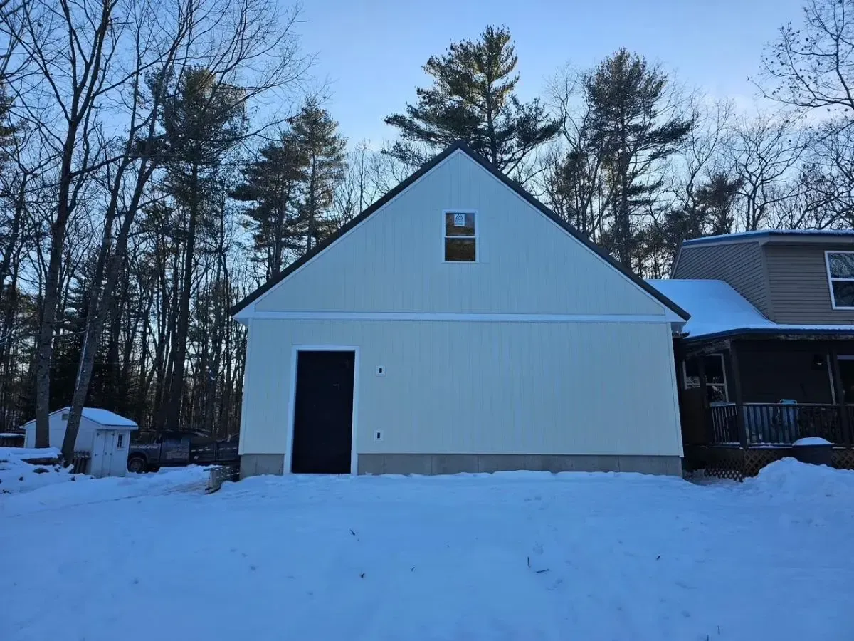 Light yellow building with a black door and window in a snowy yard, surrounded by trees.