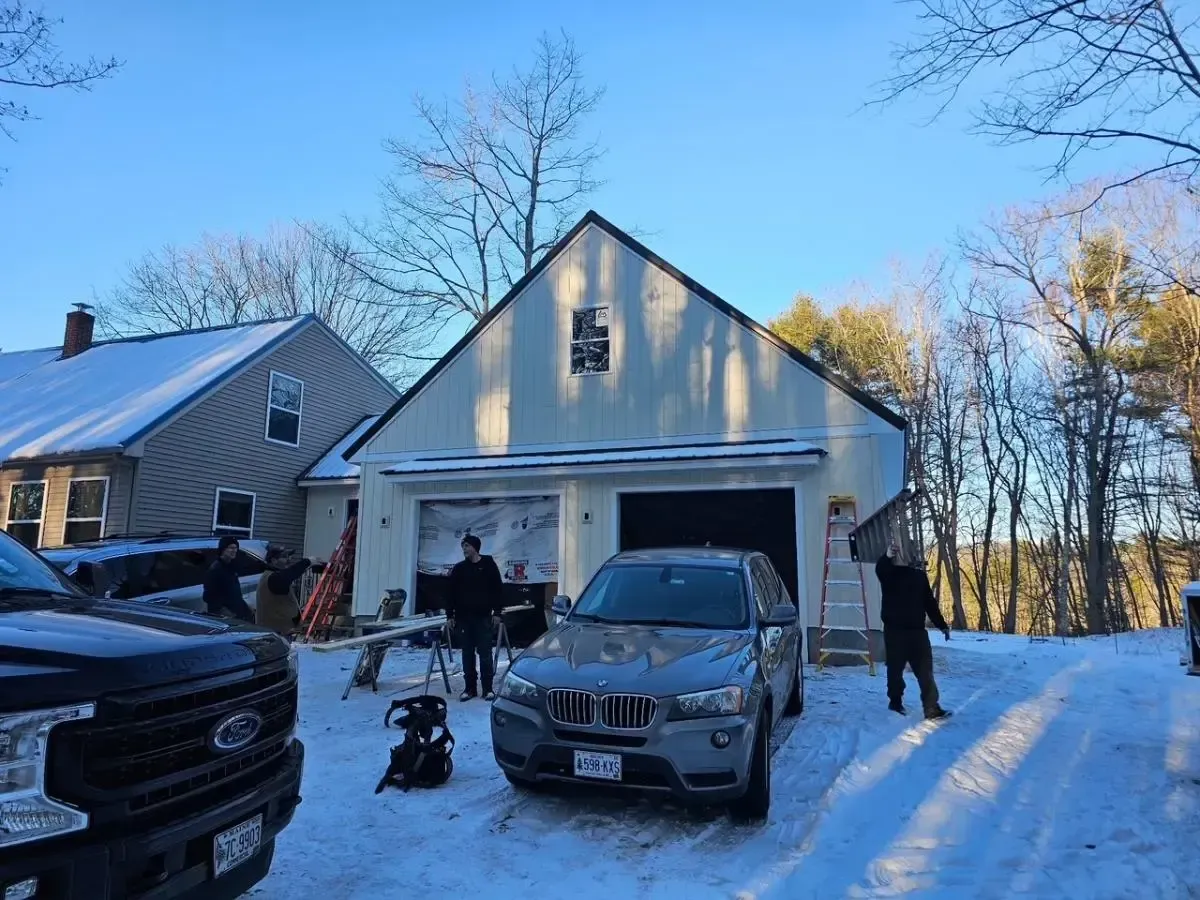 Snowy scene: Garage with open door, SUV parked, people outside working. Trees, house in background.