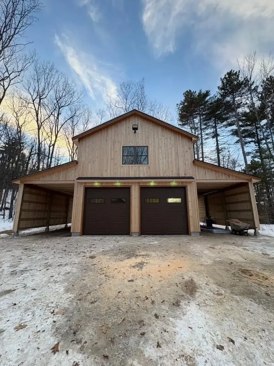 Two-story wooden barn with two garage doors and open side bays; snowy ground; trees.