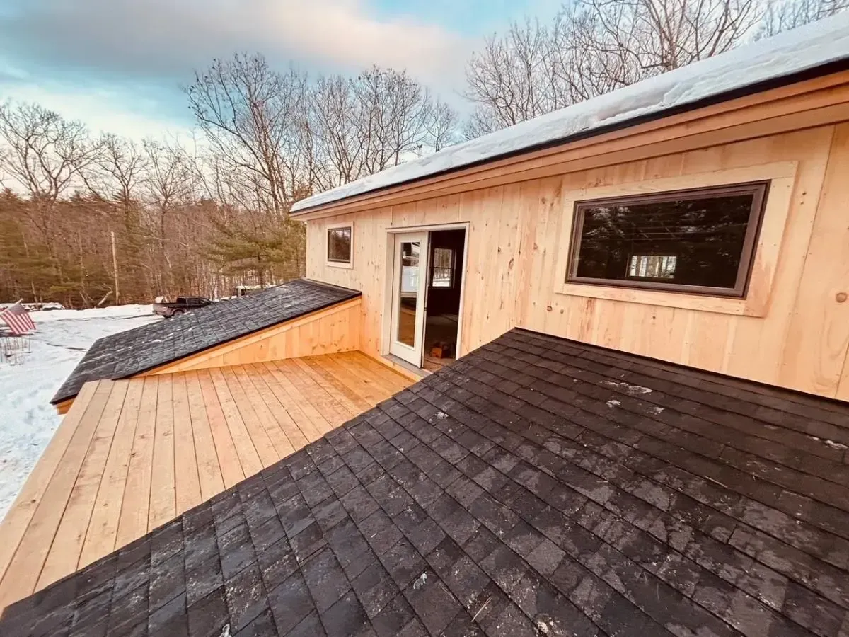 Exterior view of a house with a wooden deck and black shingled roof, set against a winter landscape with snow.