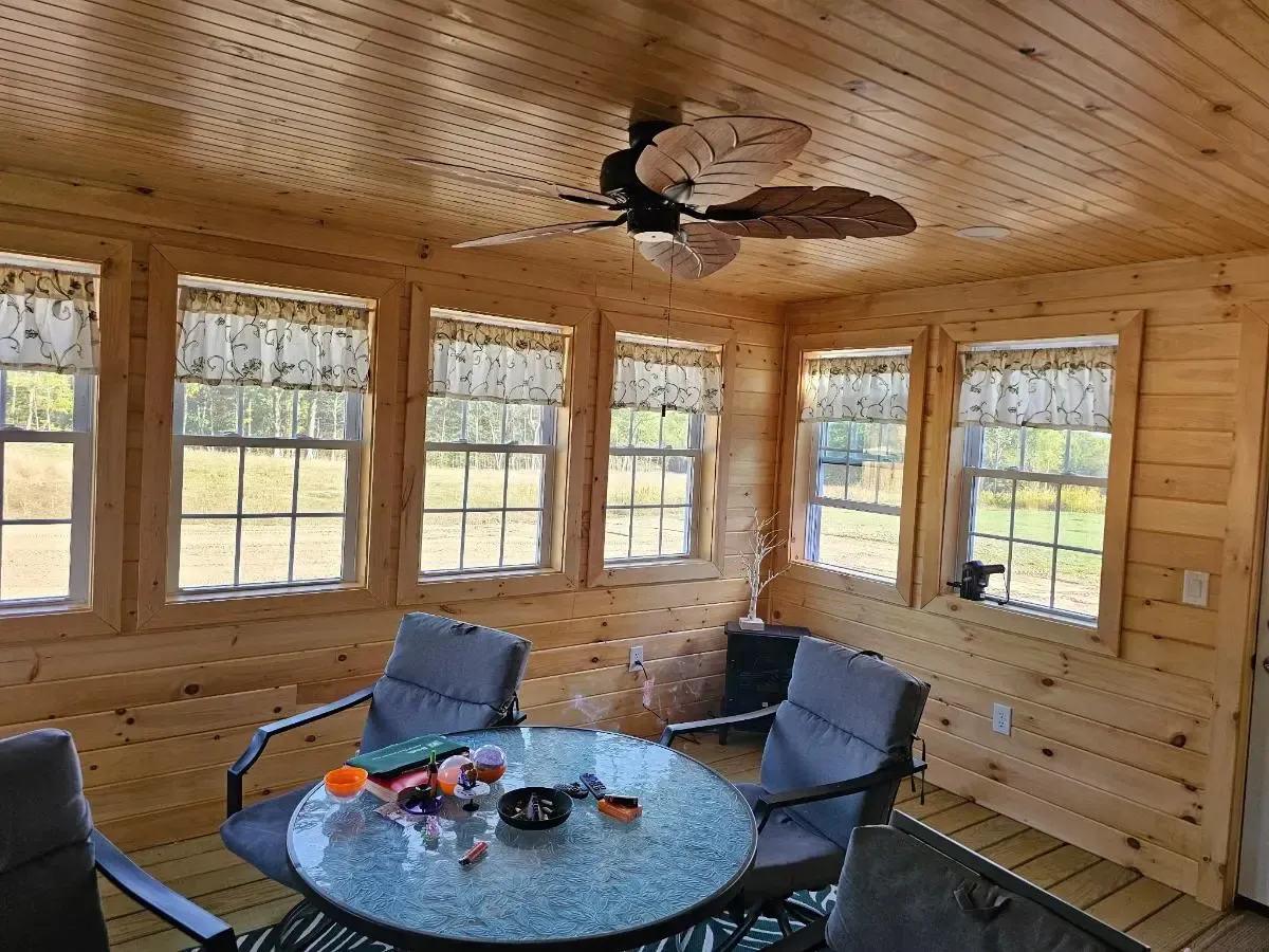 Sunroom with light wood paneling, windows, chairs around a glass table, and ceiling fan.