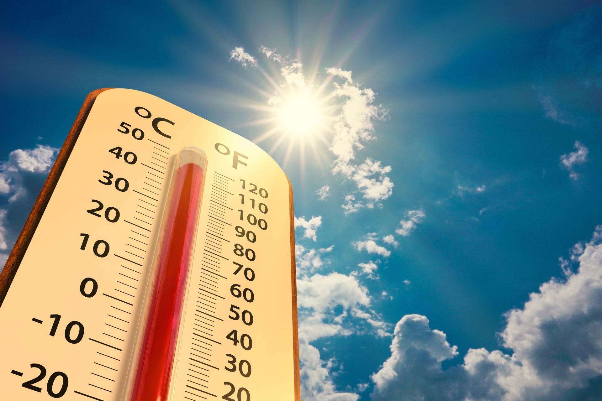 A close up of a thermometer against a blue sky with the sun shining through the clouds.