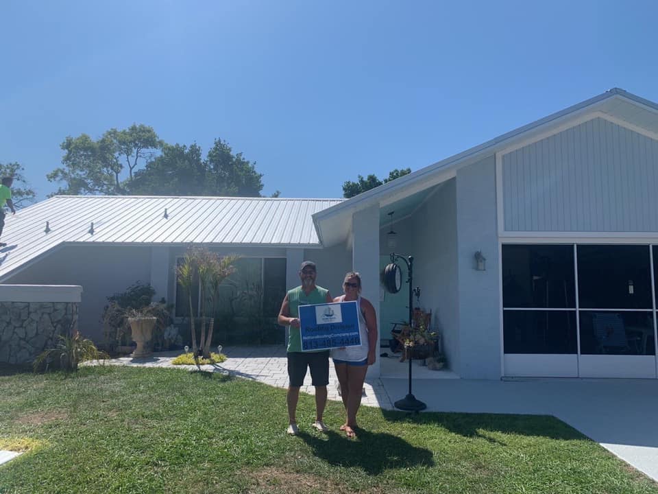 A couple stand in front of their home with its new metal roof and holding the sign for Nations R & C