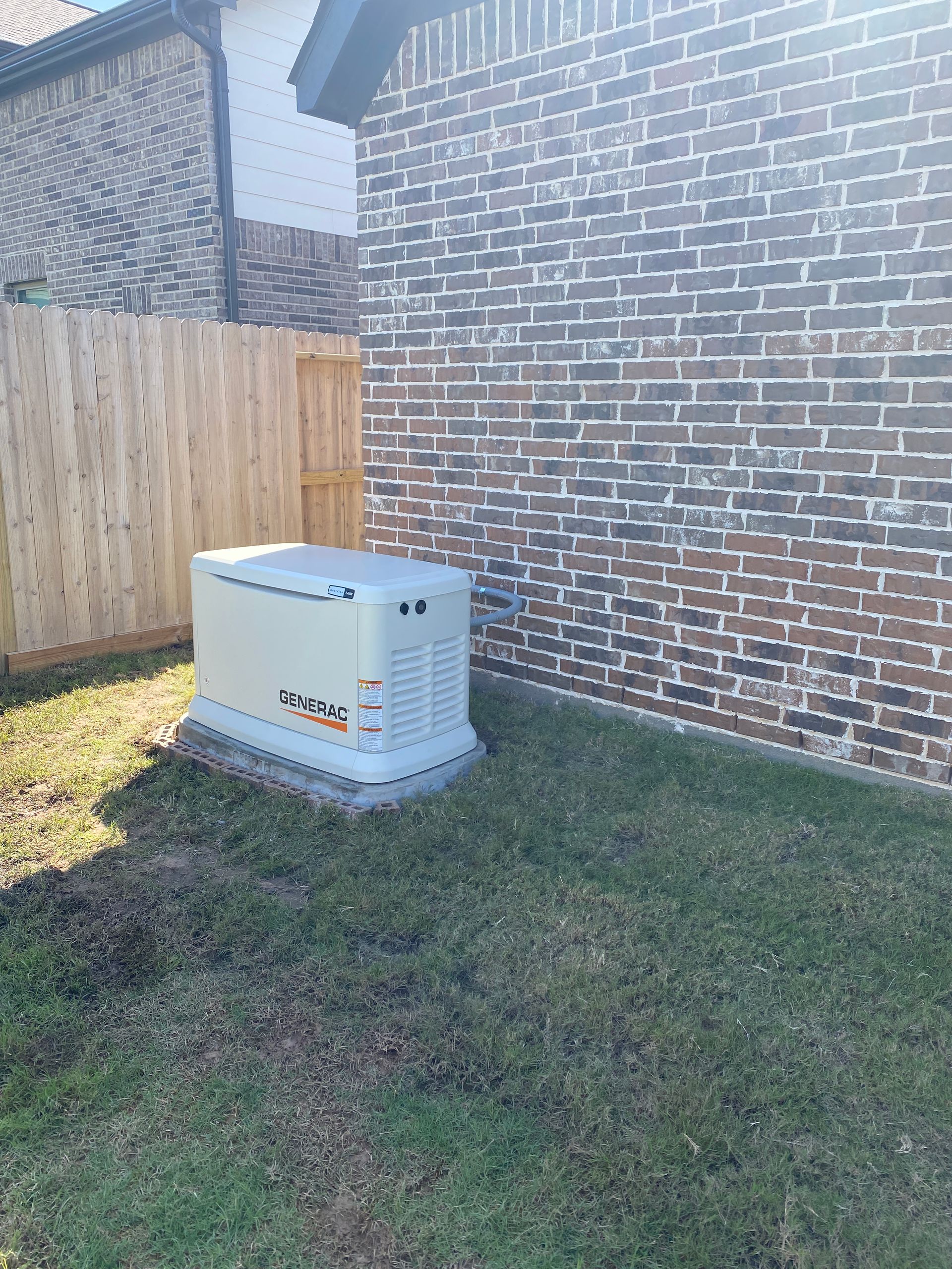 A generator is sitting in the backyard of a brick house next to a wooden fence.