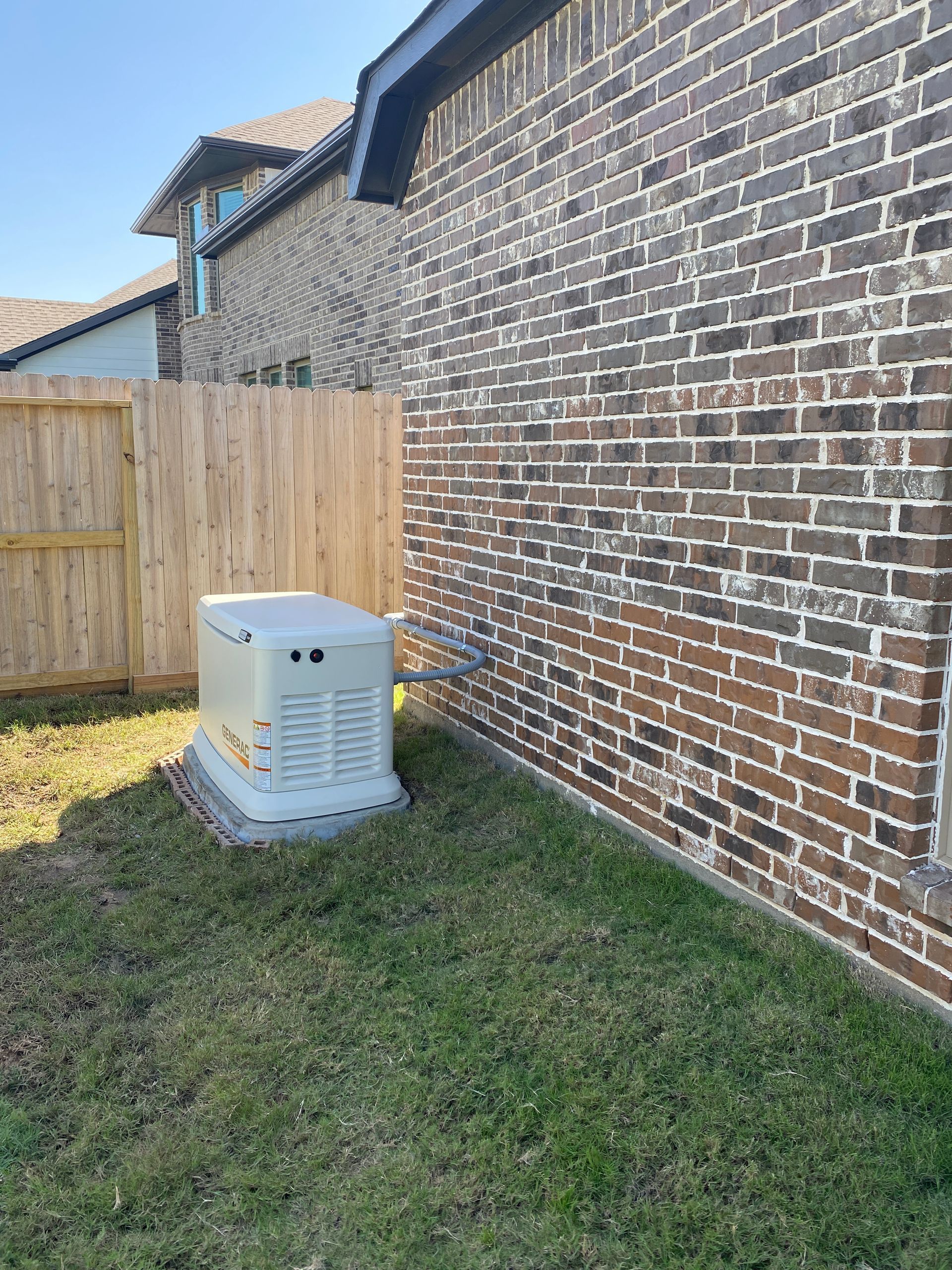 A generator is sitting on the side of a brick building next to a wooden fence.