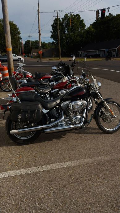 Motorcycles parked on a street near a building. Foreground has a black bike. Trees and sky in background.
