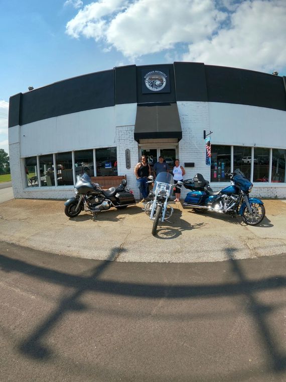 Motorcycles parked outside a white brick building with a black awning and sign. People stand at the door.