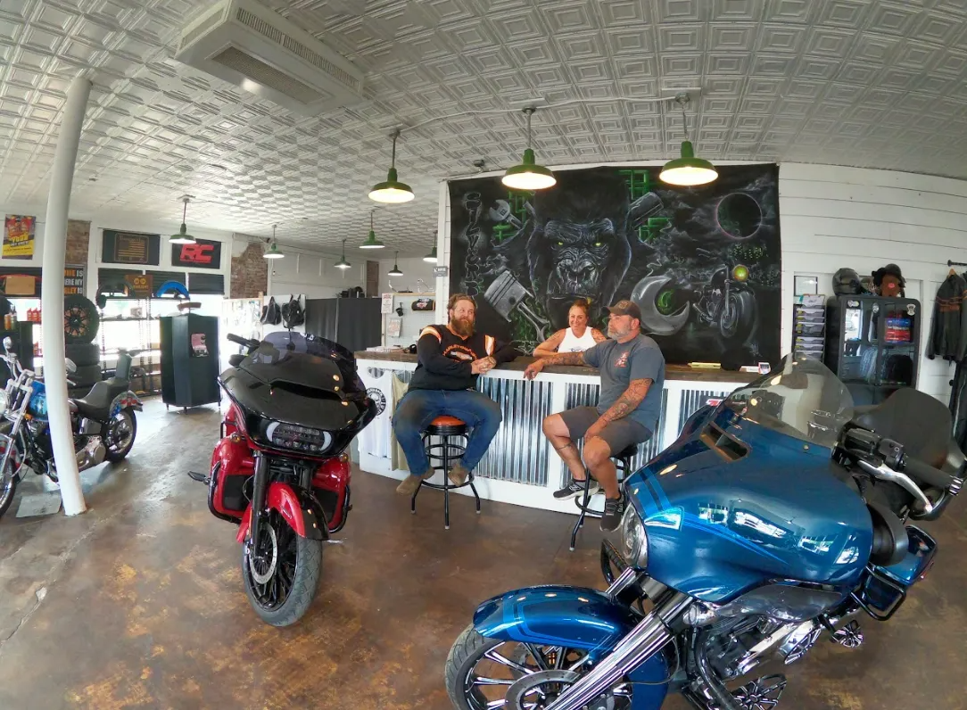 Inside a motorcycle shop, customers and employees seated at a counter next to a chalkboard art piece. Two motorcycles in the foreground.