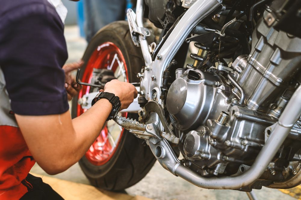 Mechanic repairing a motorcycle engine; close-up view with red wheel, gray metal, and tool in hand.