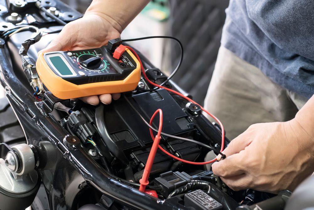 Person using a multimeter to test motorcycle's electrical system, orange meter, black motorcycle, hands.
