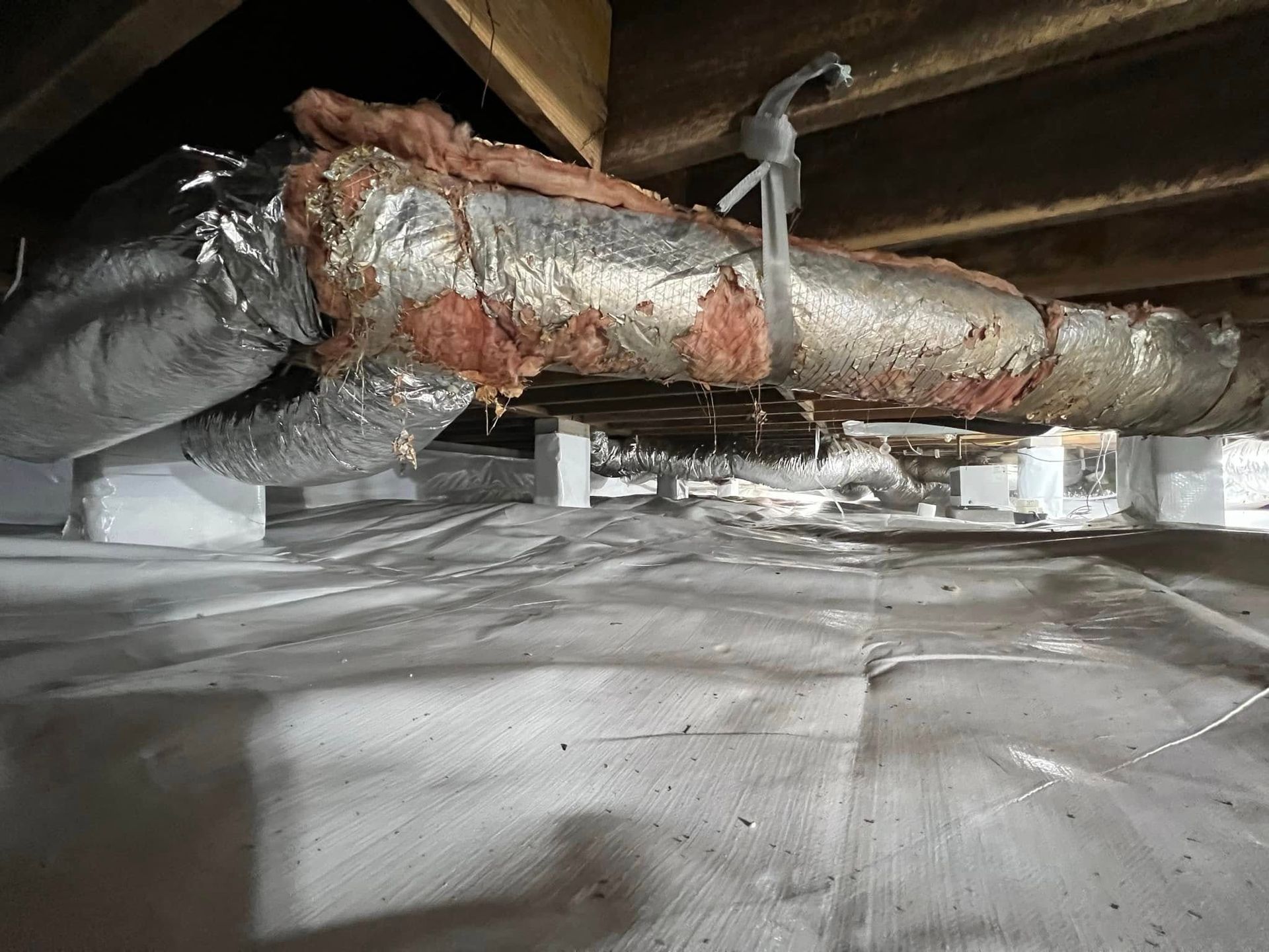 Under-house view with ductwork and insulation. White supports and vapor barrier cover the ground.