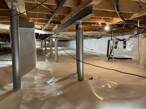 Crawl space with supports, white plastic sheeting, electrical wiring, and insulation against a white wall.