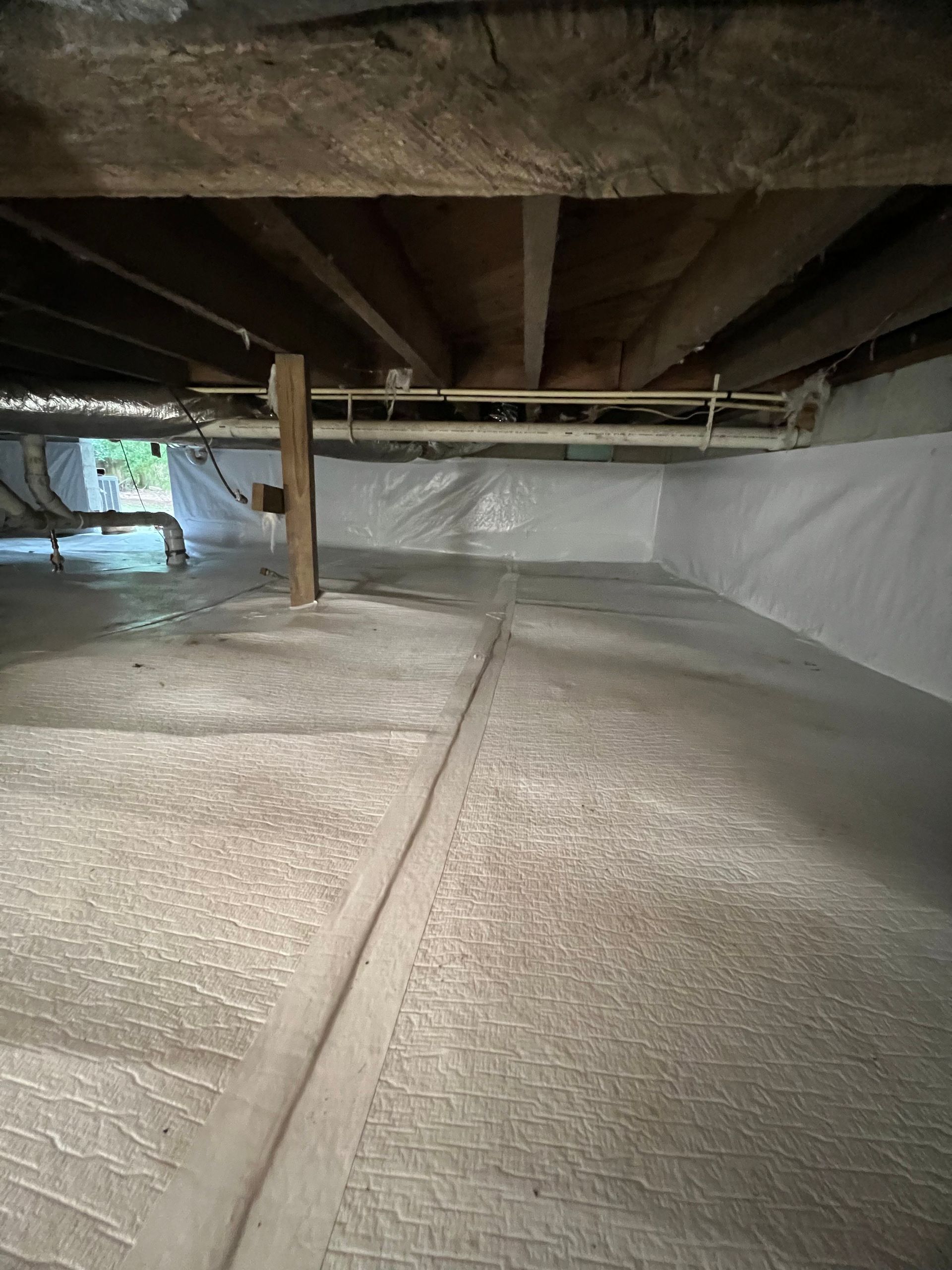 Crawlspace with insulation on walls and floor, beams above, pipes, and wooden support beams.