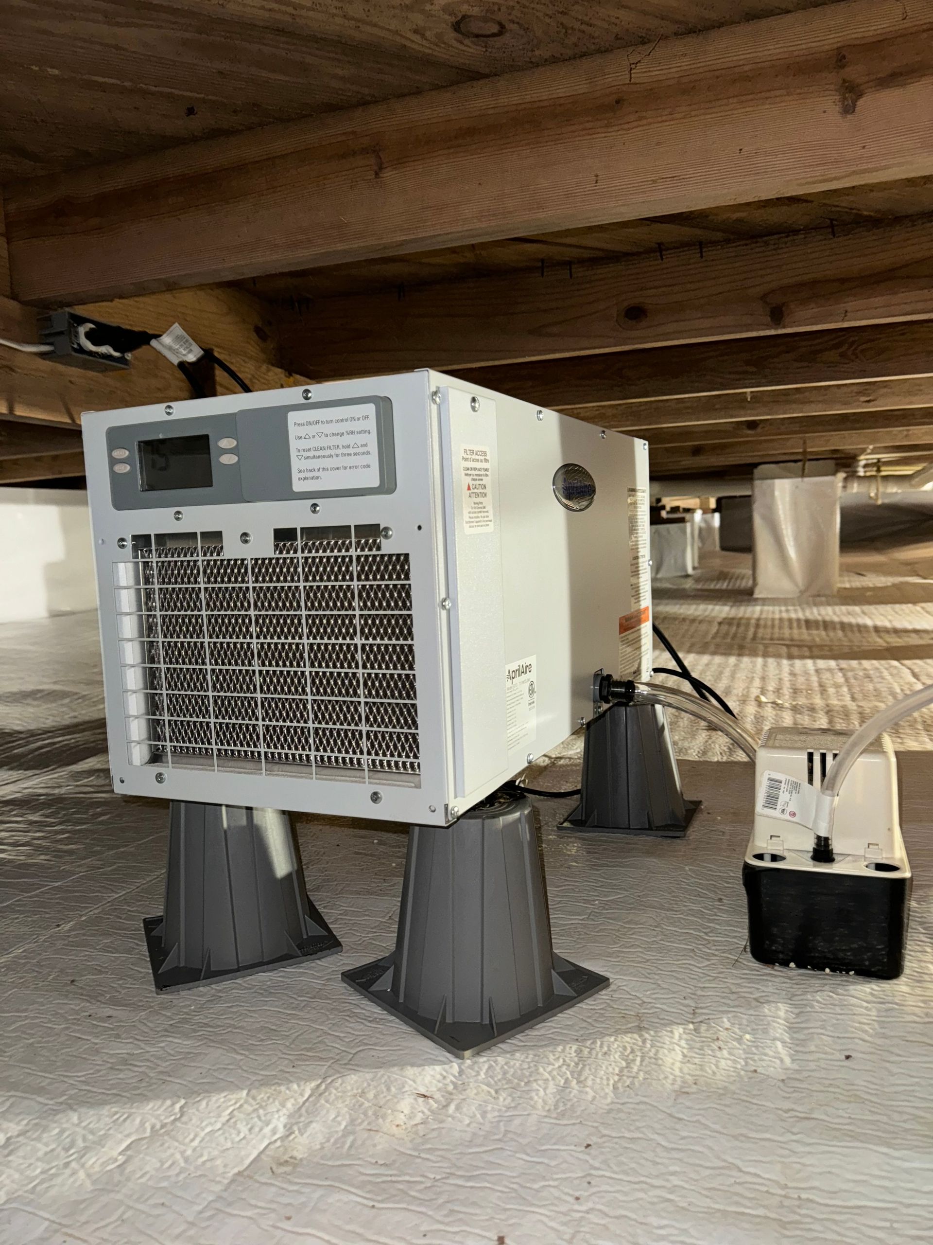 Crawl space with a dehumidifier on blocks, pump, and plastic vapor barrier. Wooden beams above.