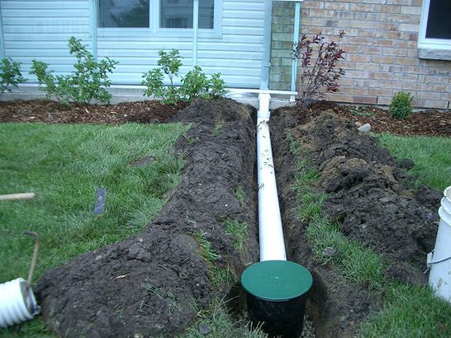 A trench with white drainage pipes being installed near a house. Earth and a green lid are visible.