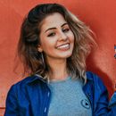 A woman wearing a denim jacket and a grey shirt is smiling in front of a red wall.