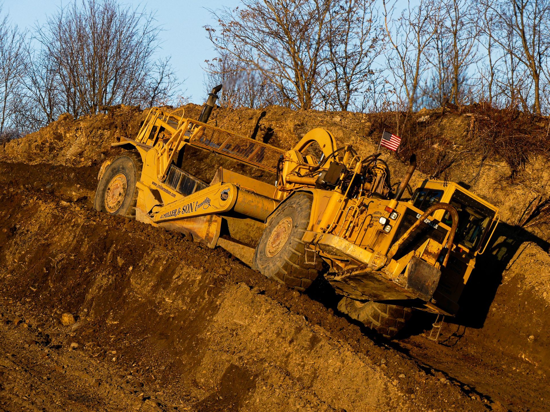 Yellow earthmover on a dirt pile, angled upward. Brown soil, blue sky, trees in the background.