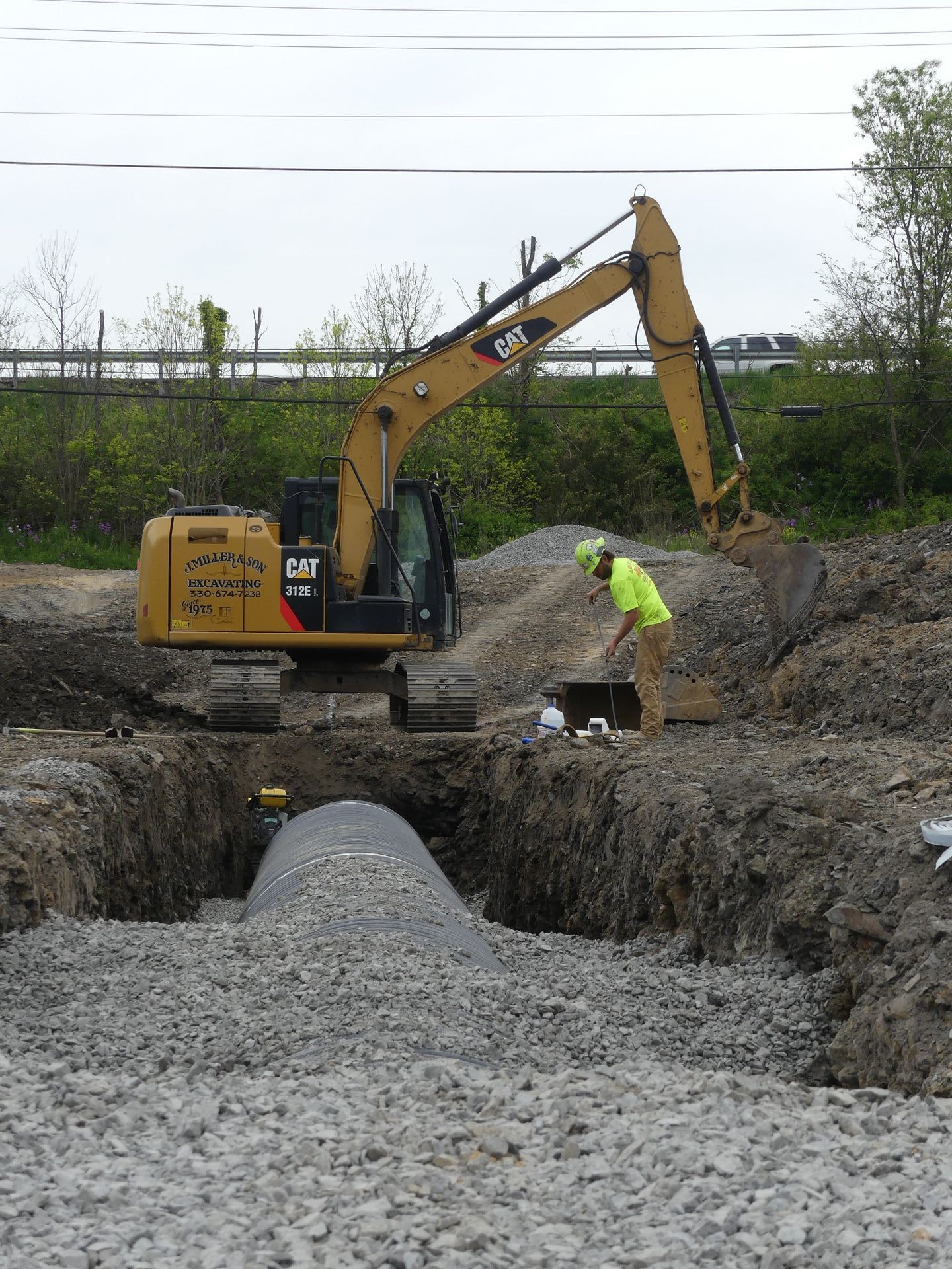 Construction site: Excavator and worker near gravel-filled trench.