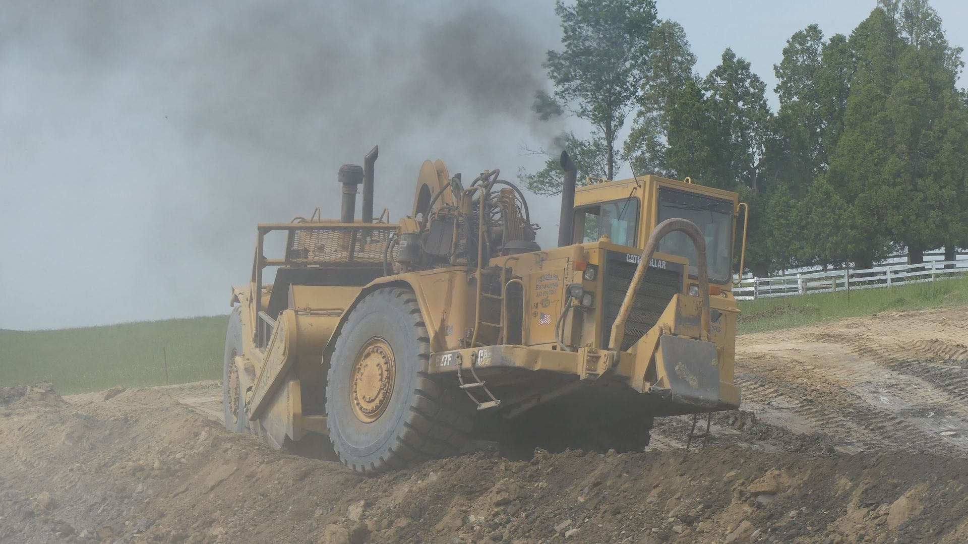 Yellow earthmover kicking up dust on a dirt hill near trees and a white fence.