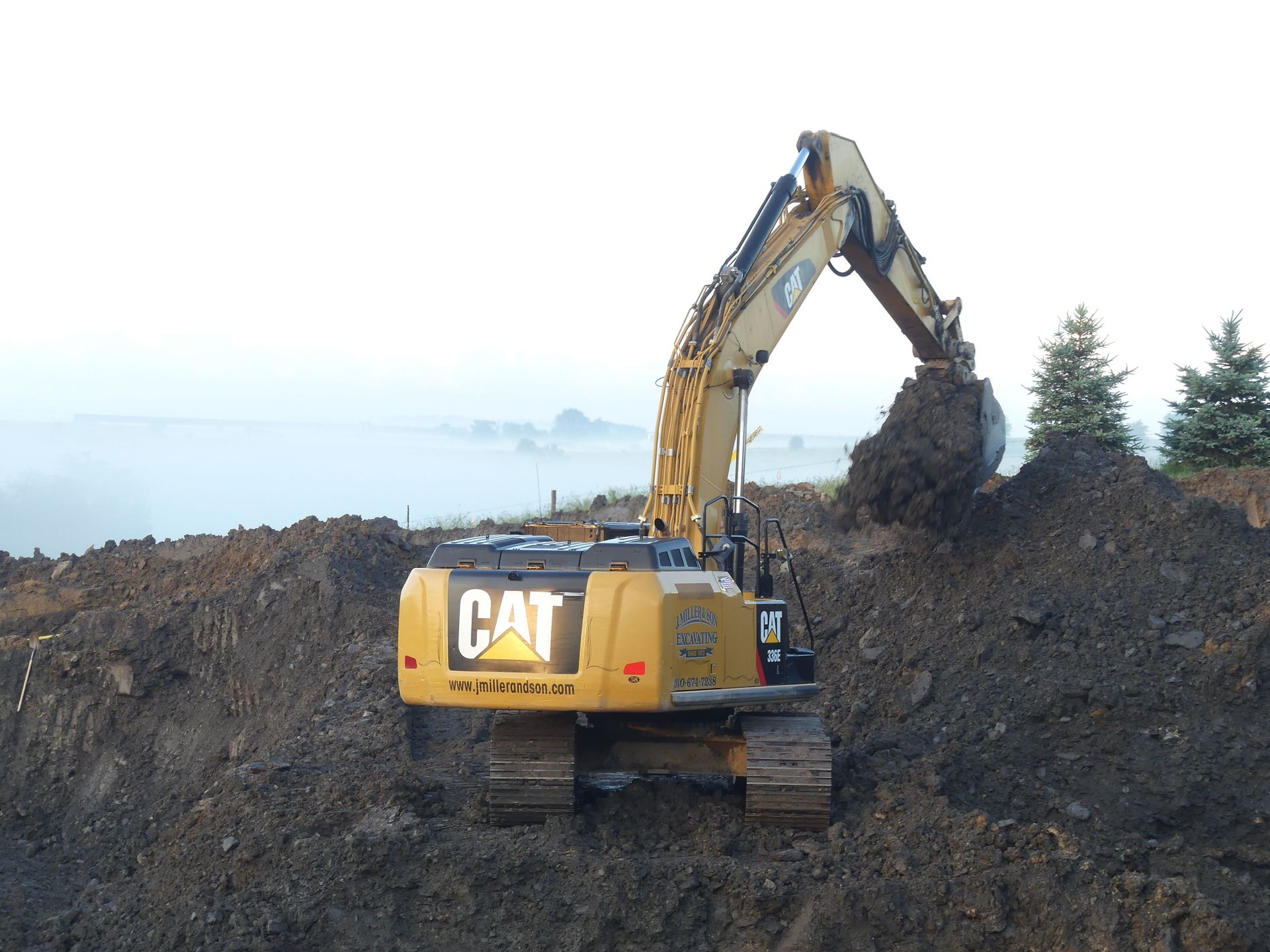 Yellow Caterpillar excavator digging in a dirt pile under a hazy sky.