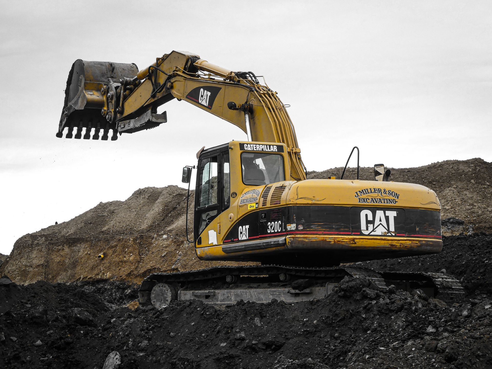 Yellow Caterpillar excavator digging in a dark dirt pile against a cloudy sky.