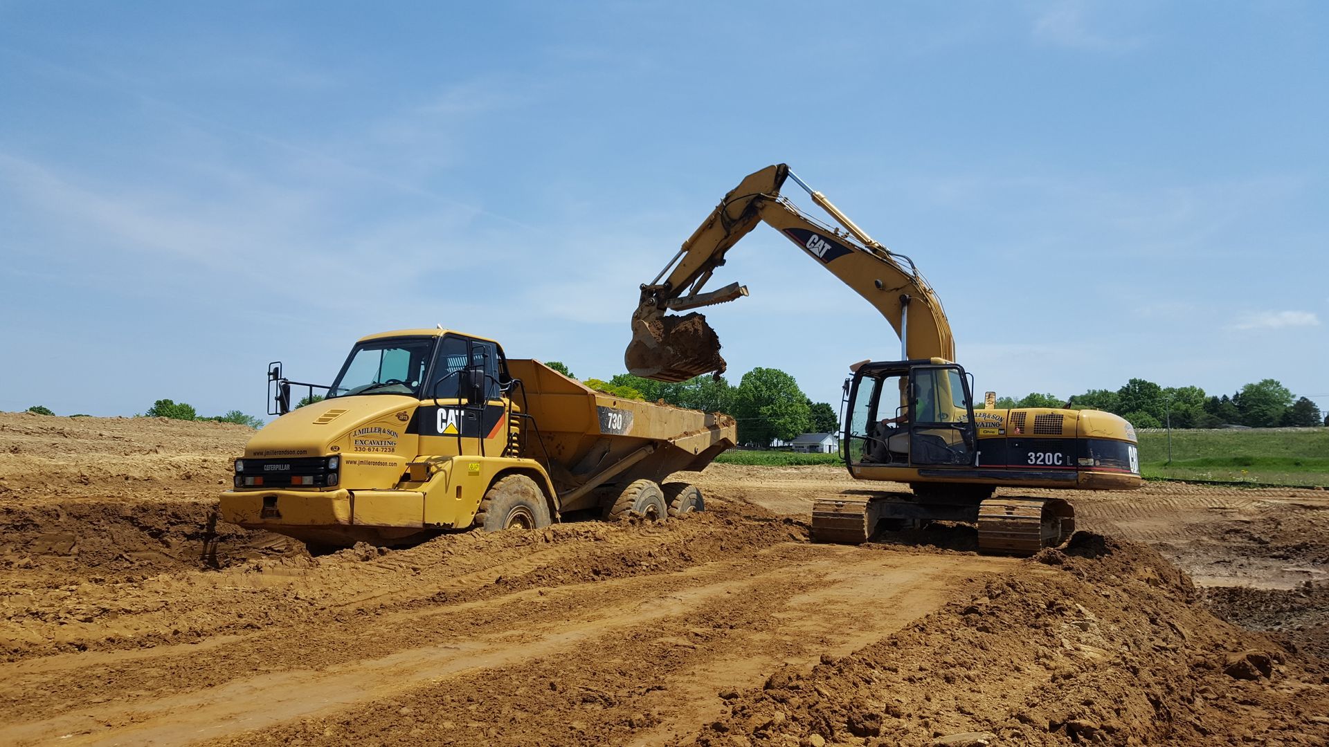 An excavator loads dirt into a dump truck on a muddy construction site under a blue sky.