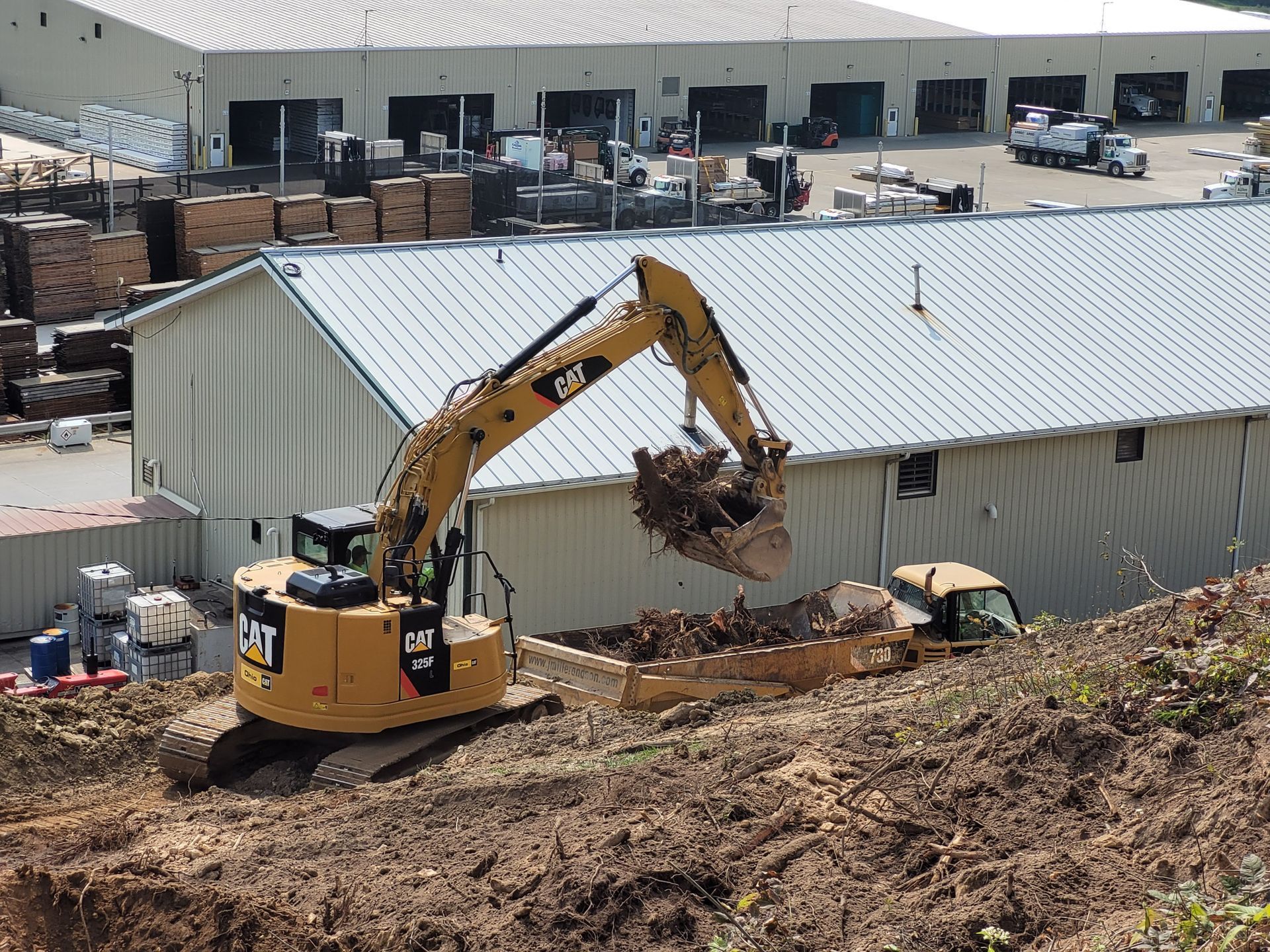 Yellow Caterpillar excavator loading dirt near industrial buildings.