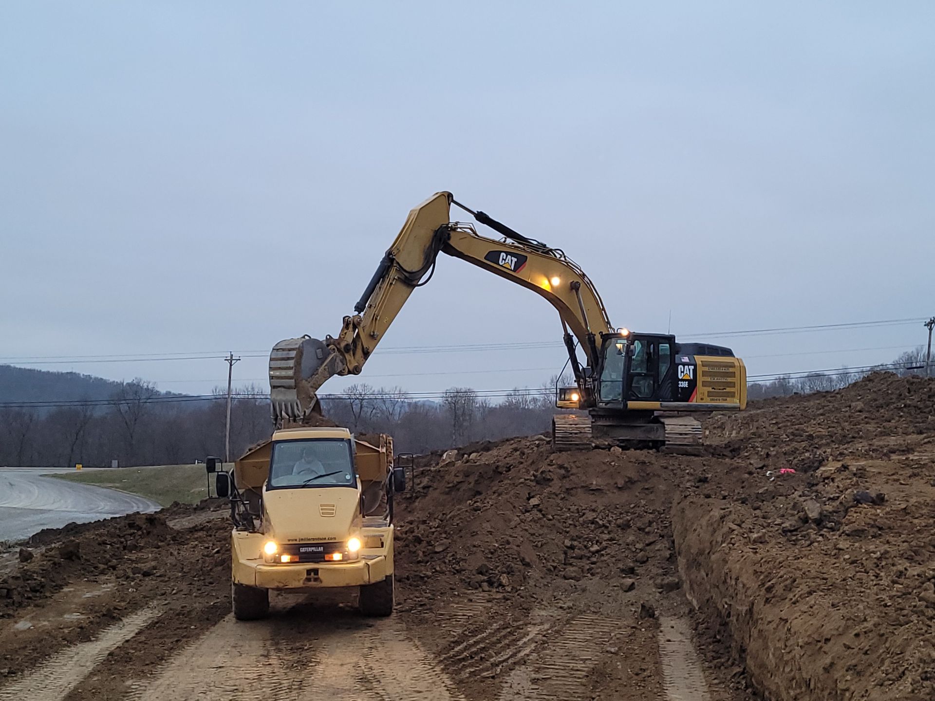 Two yellow construction vehicles on a muddy embankment, one excavating, the other in the foreground. Overcast sky.