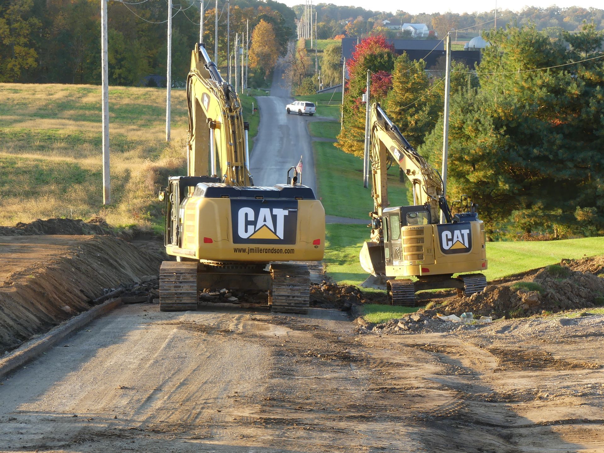 Two yellow CAT excavators on a road, digging. A white car drives in the distance on the uphill road.