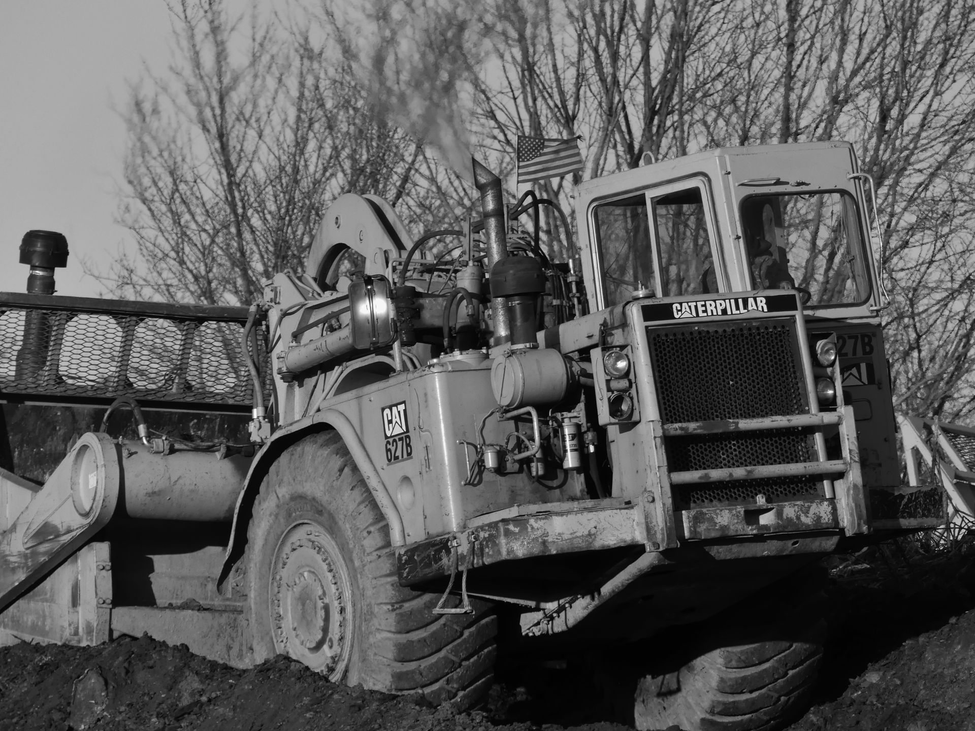 A large Caterpillar earthmover stuck in mud, with an American flag attached, set against a backdrop of bare trees.