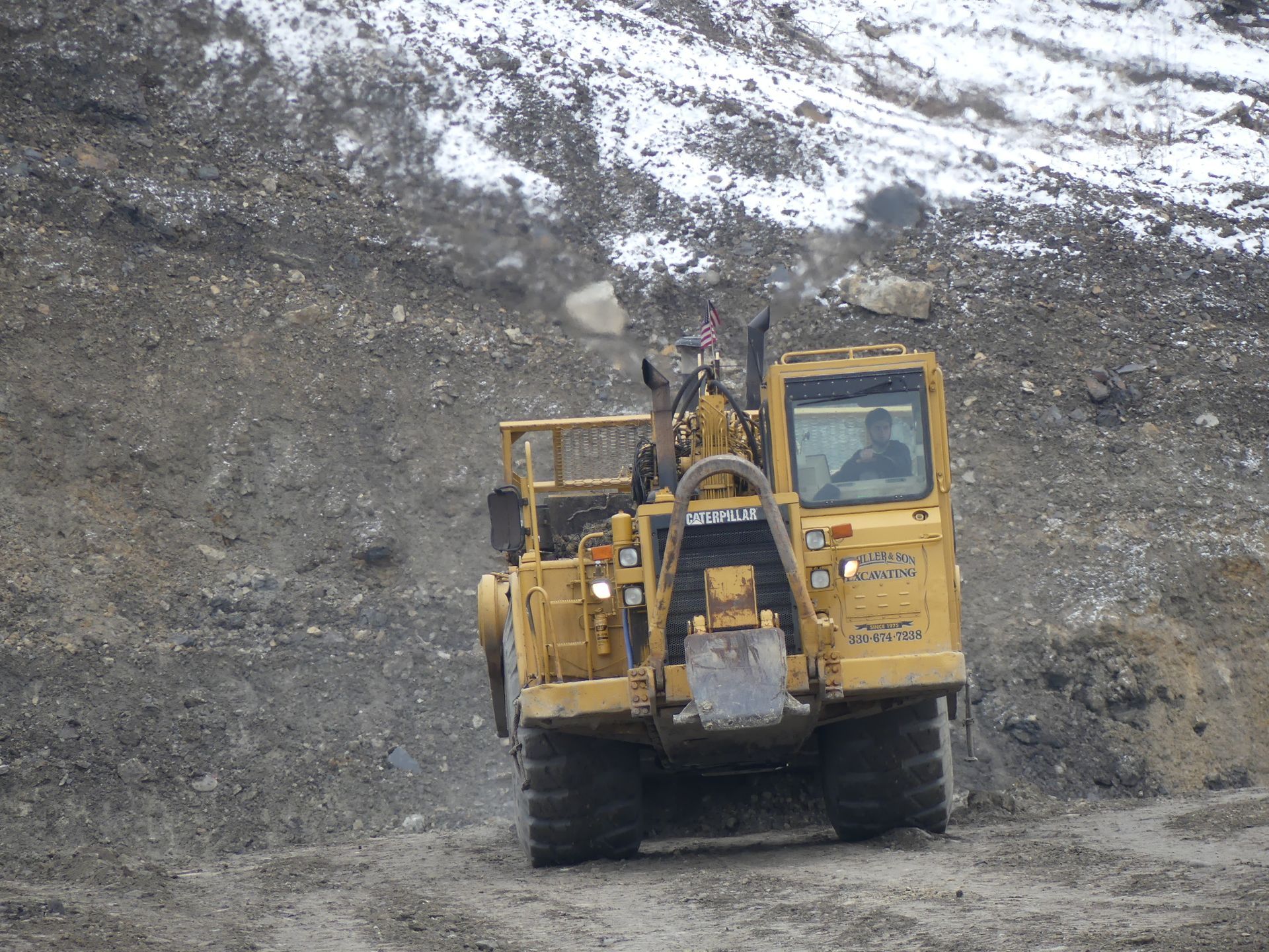 Yellow construction vehicle driving on a gravel surface near a snowy hillside.