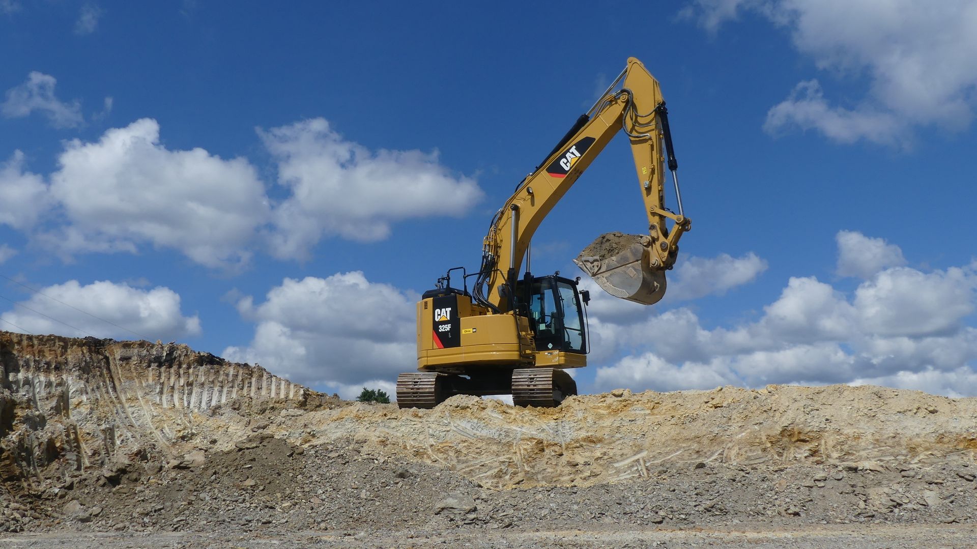 Yellow excavator digging on a dirt mound under a blue sky with fluffy white clouds.