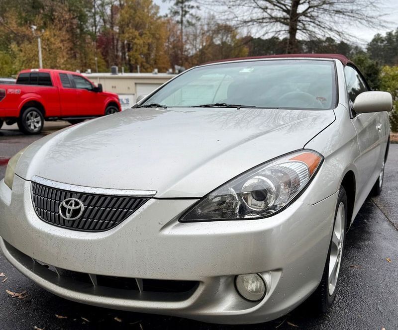 A silver Toyota Solara convertible parked outdoors on a wet surface with a red pickup truck in the background.