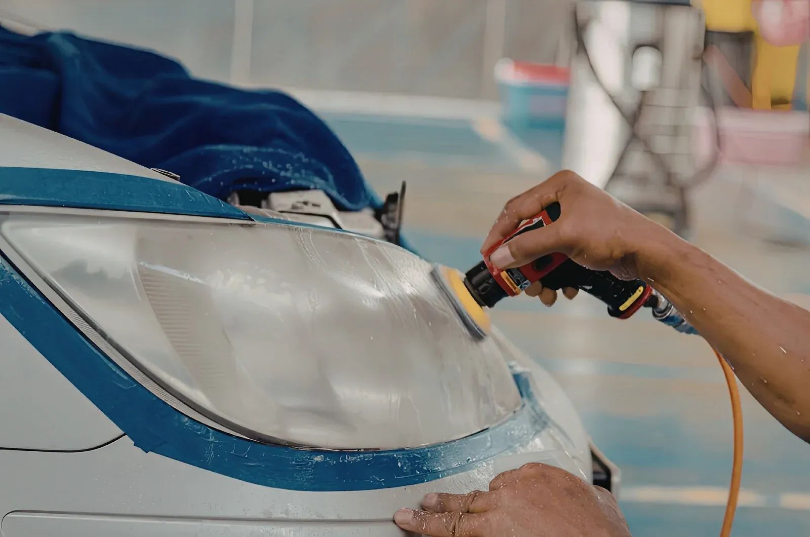 Person applies polish to a white car’s headlight with a power tool in a workshop.