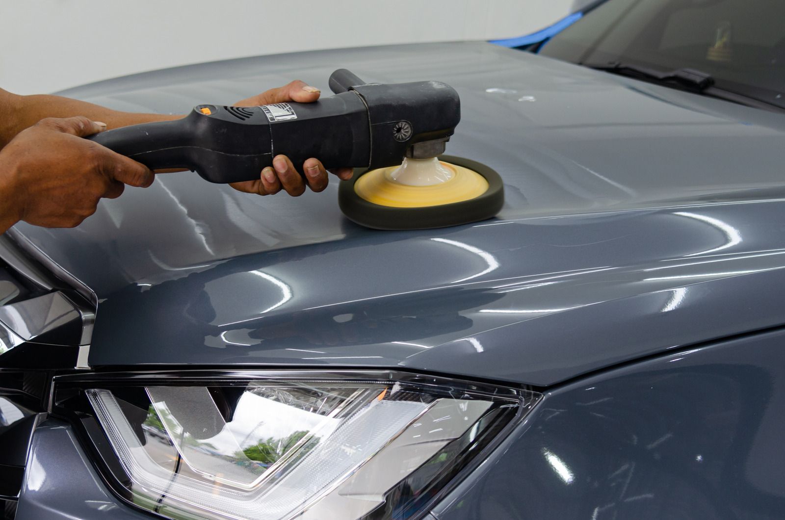 A person uses a rotary buffer tool to polish the hood of a gray car.