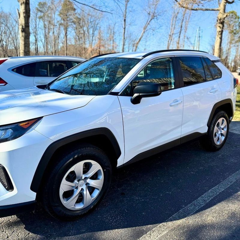 A white Toyota RAV4 parked on an asphalt lot with a treeline in the background under a clear sky.