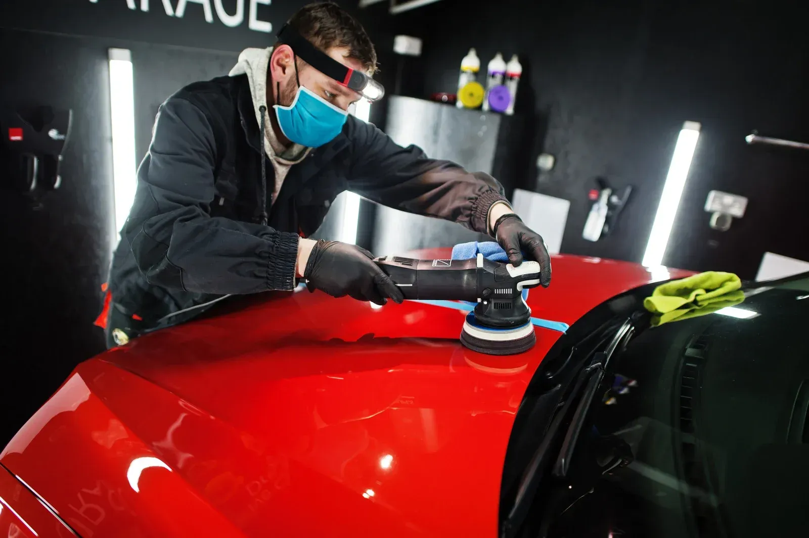 A professional technician in a garage polishes the hood of a red car using a mechanical buffer.