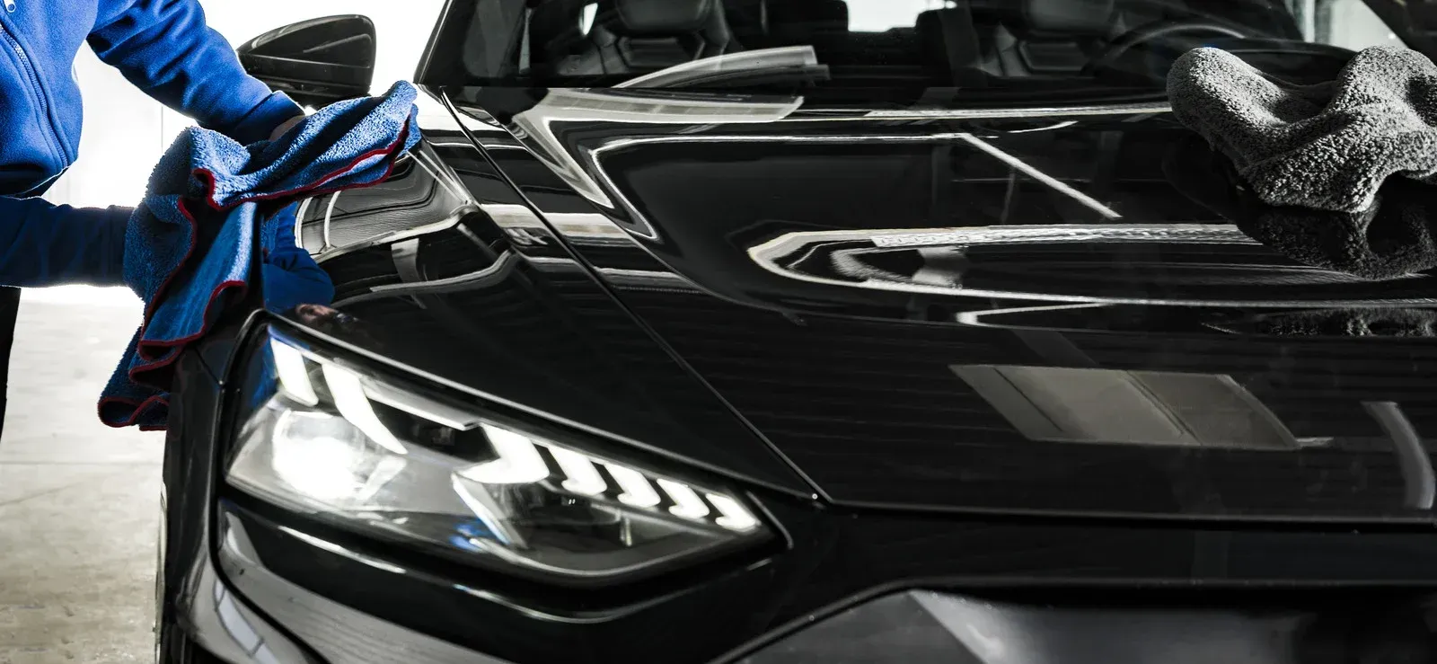 A person in a blue shirt uses a cloth to wipe down the glossy black hood and headlight of a sports car.