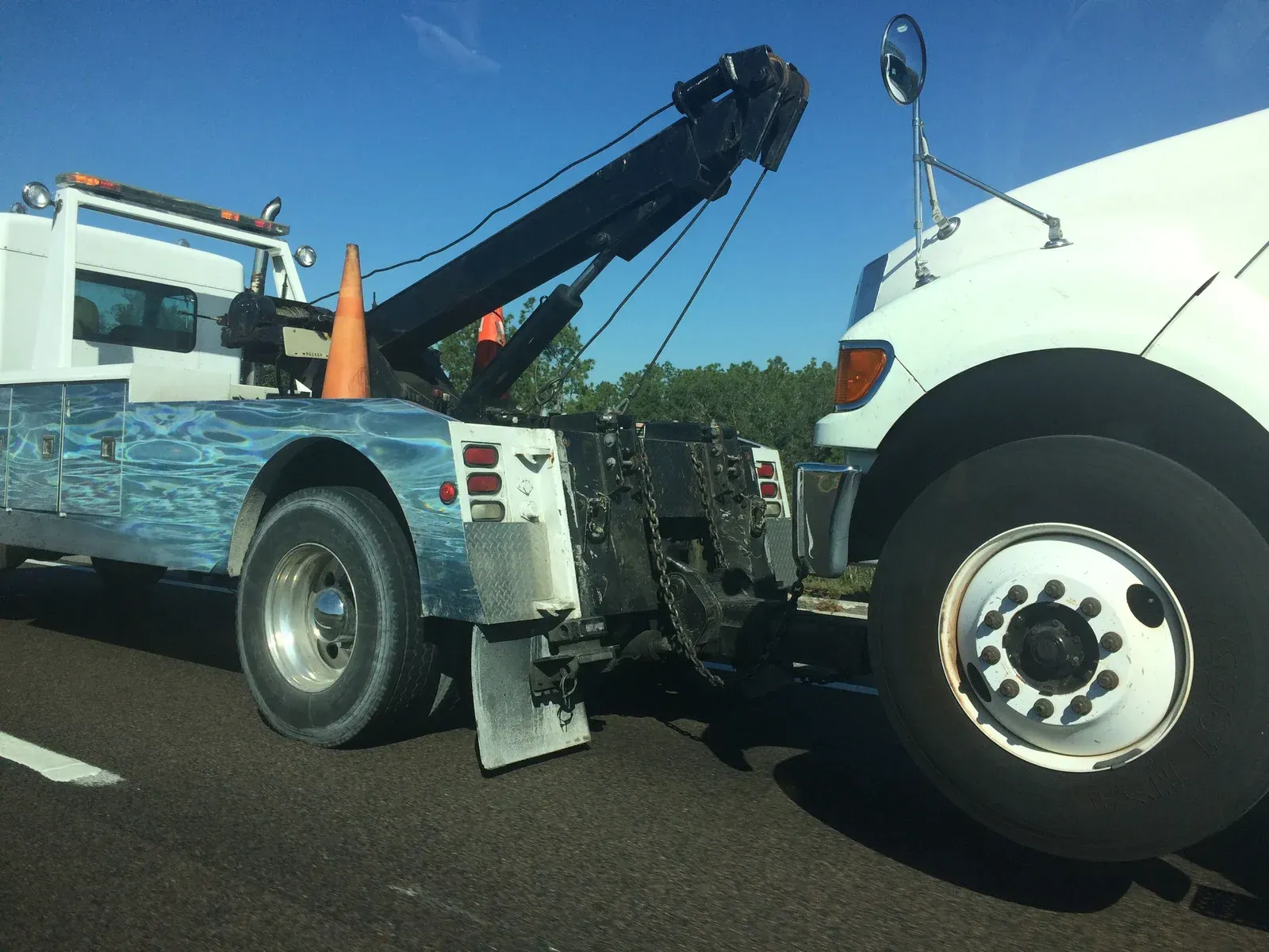A white tow truck is towing a large white commercial semi-truck on a road under a clear blue sky.