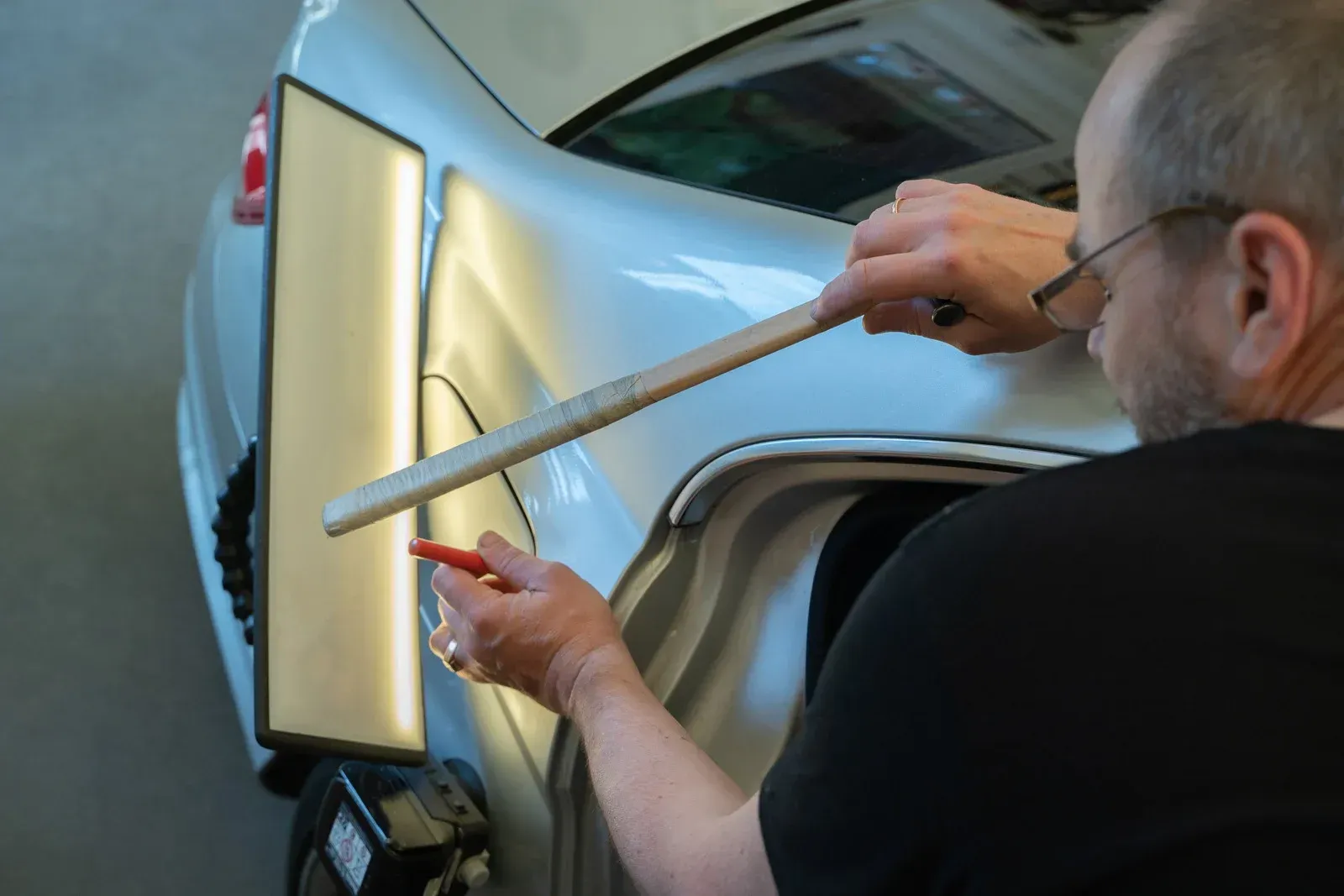 A technician uses a light board and a metal tool to perform paintless dent repair on a silver car's rear quarter panel.